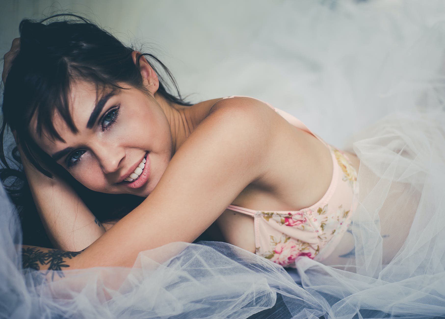 close up photo of smiling woman wearing pink floral brassiere lying on white sheer cloth