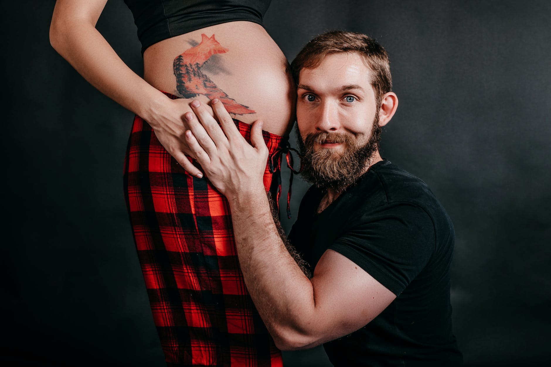 hipster man embracing faceless pregnant partner on black background